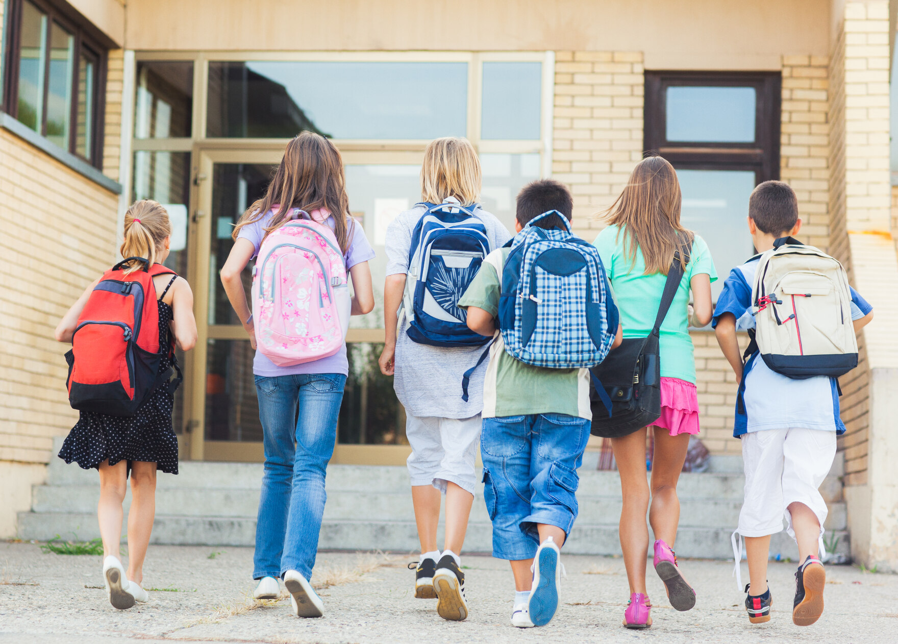 Sechs Kinder gehen Hand in Hand mit Rucksack auf dem Rücken in RIchtung einer Schule