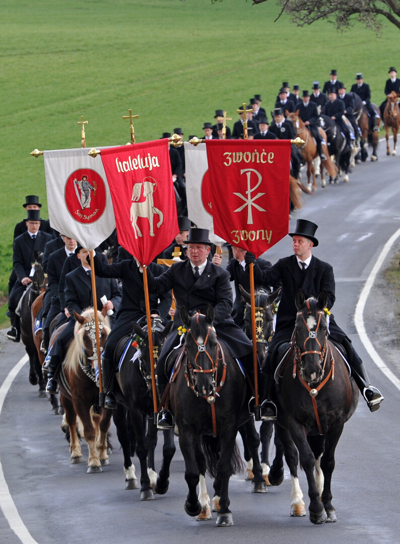 Eine große Gruppe Reiter in festlichem Gewand reiten auf einer Straße.