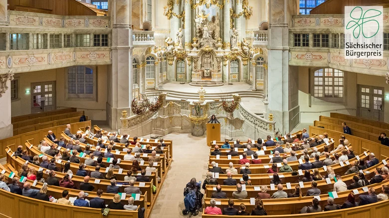 Männer und Frauen sitzen in einer Kirch. Vor Ihnen steht eine Frau an einem Rednerpult.