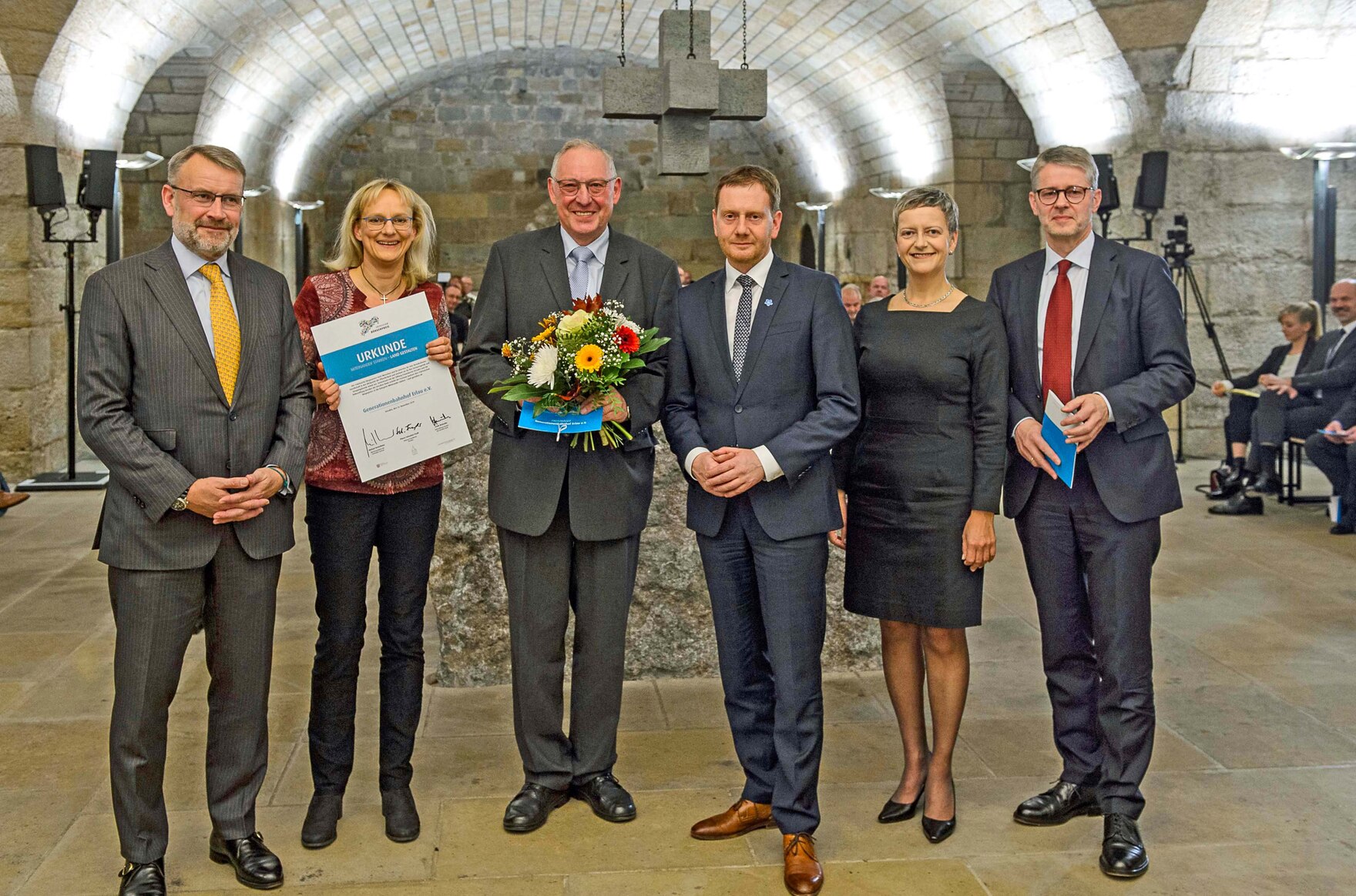 Männer und Frauen stehen in einem Gewölbe in einer Reihe. EIne Frau hält eine Urkunde in der Hand, ein Mann einen Blumenstrauß.