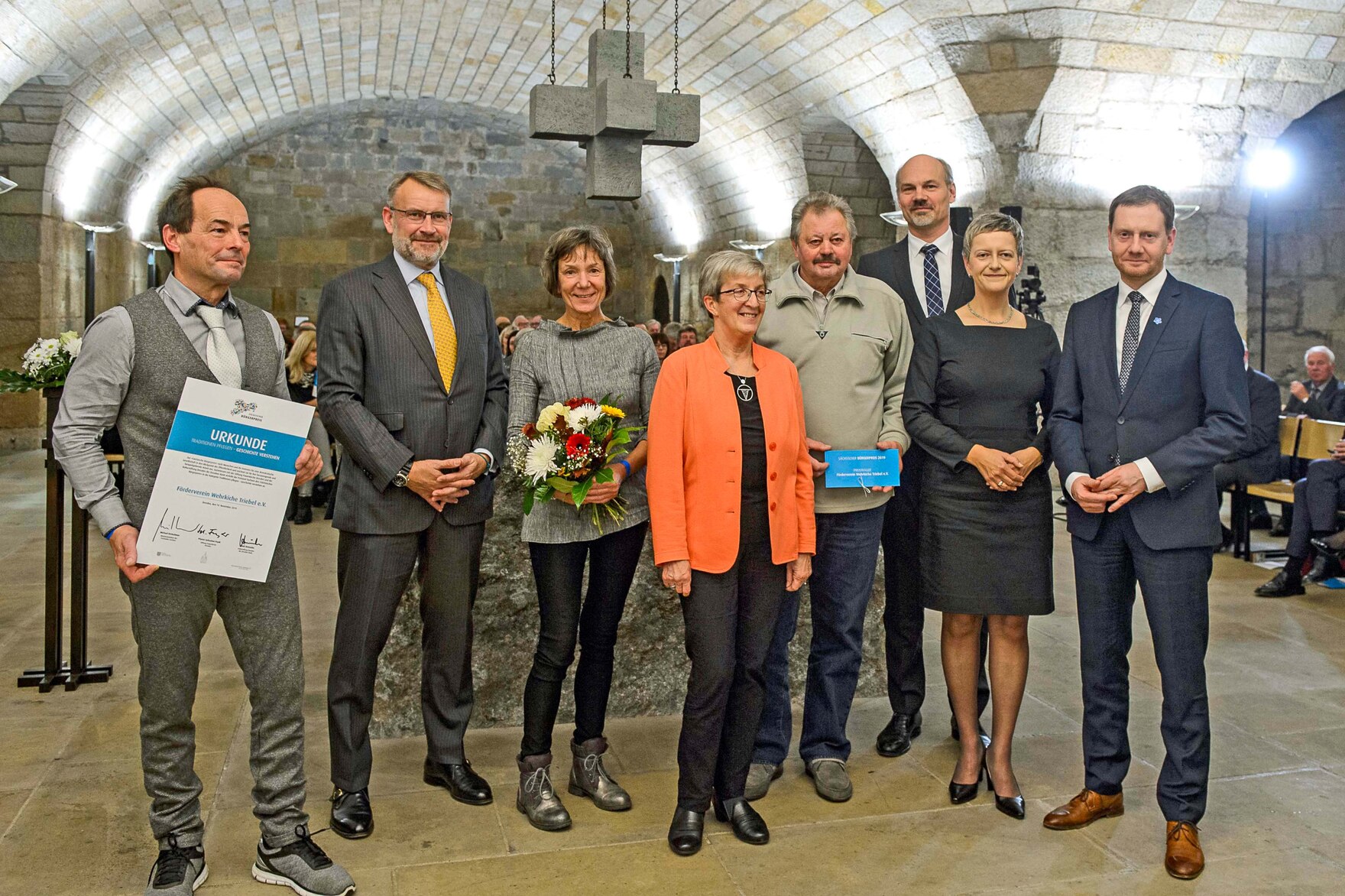 Männer und Frauen stehen in einem Gewölbe. Ein Mann hält eine Urkunde in der Hand, eine der Frauen einen Blumenstrauß.