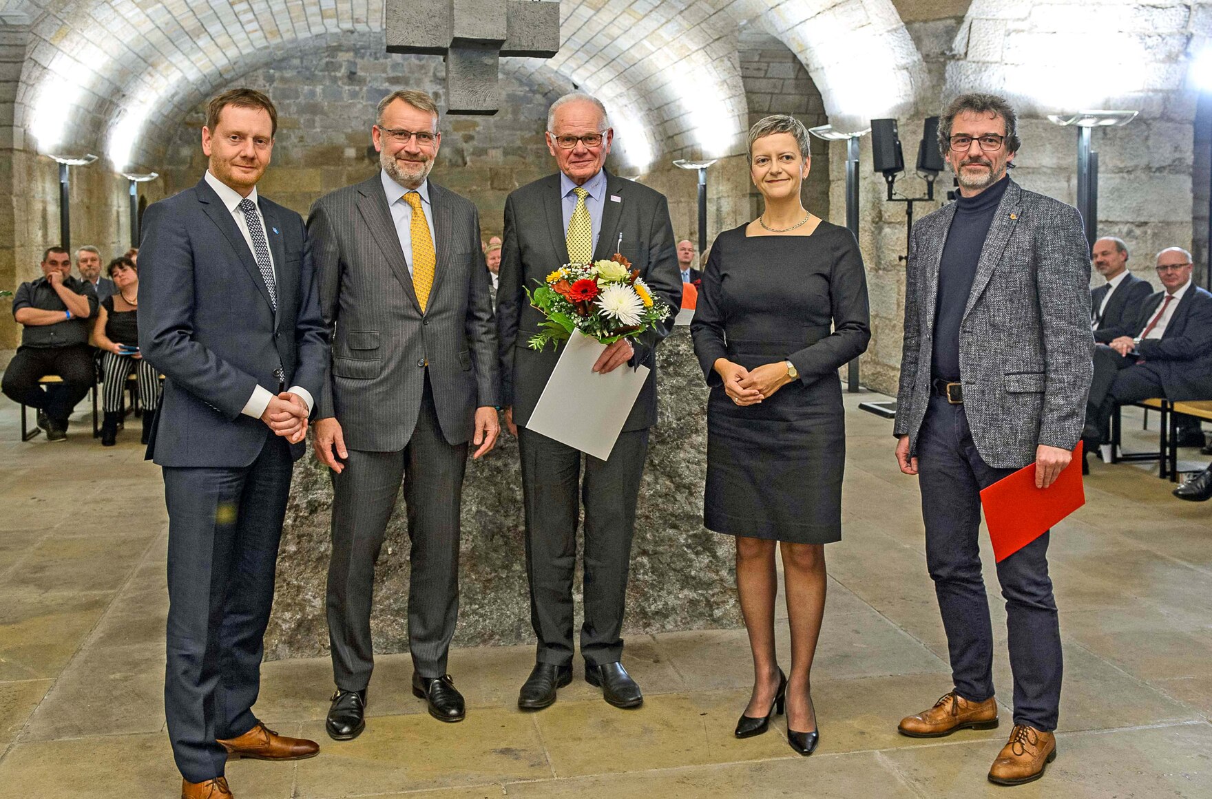 Männer und Frauen stehen in einem Gewölbe. Ein Mann hölt einen Blumenstrauß in der Hand.