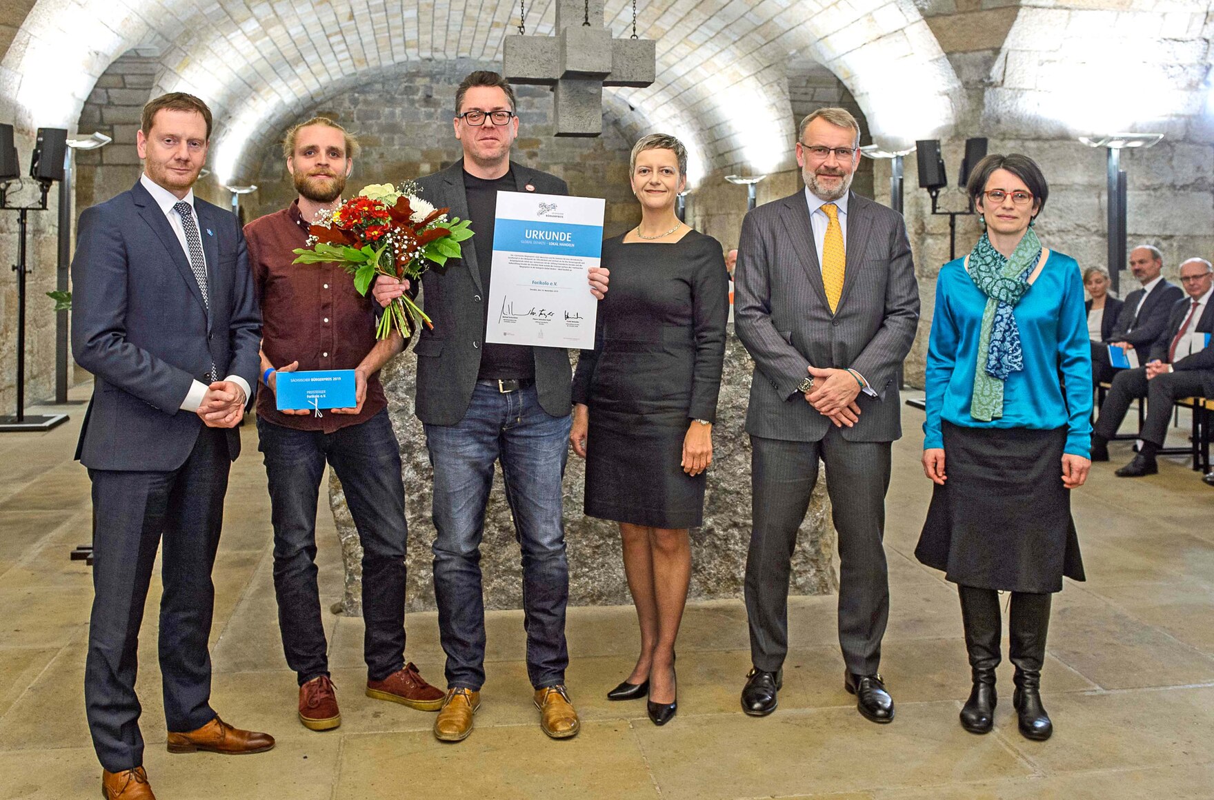 Männer und Frauen stehen in einem Gewölbe. Ein Mann hält eine Urkunde in der Hand.