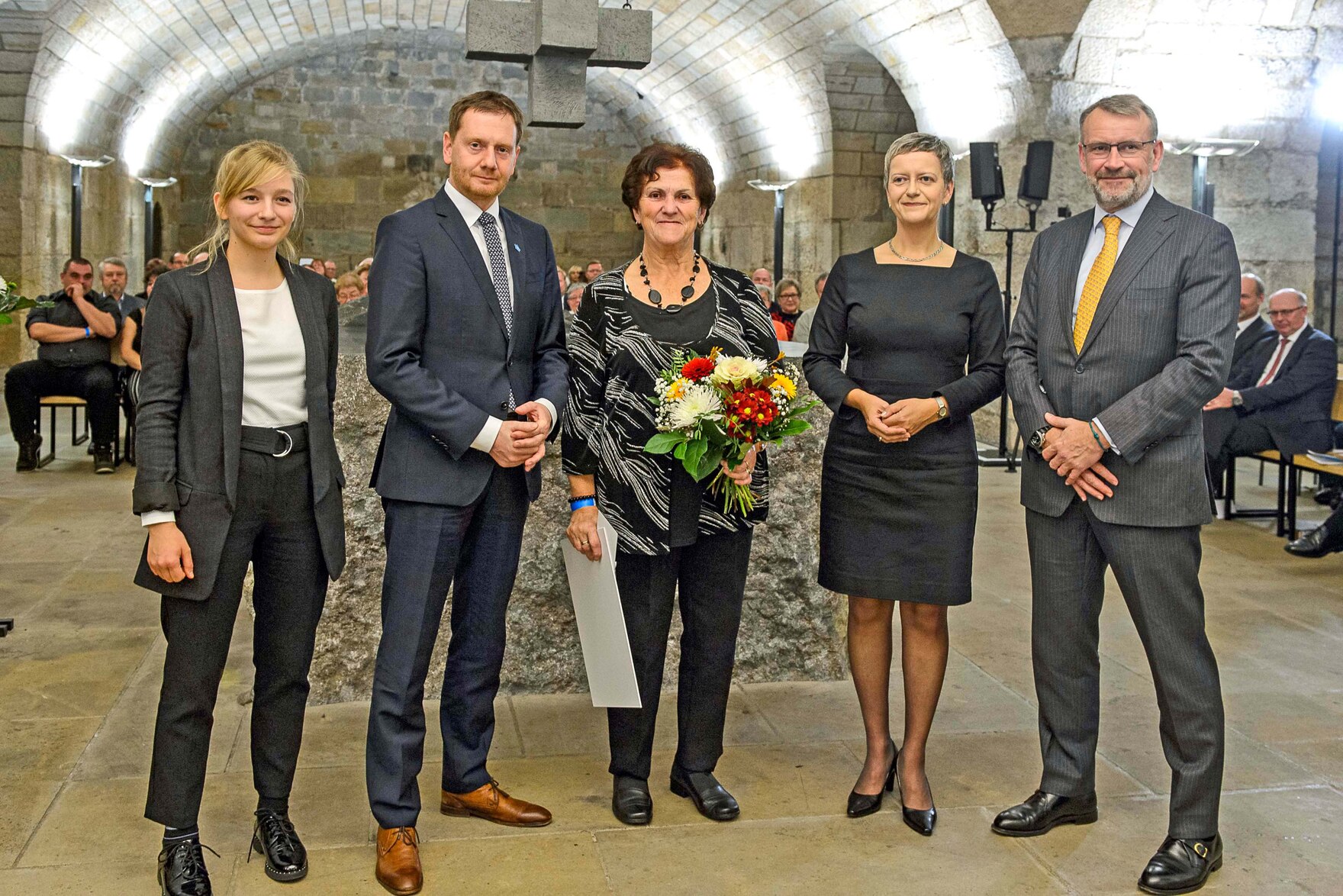 Männer und Frauen stehen in einem Gewölbe. Eine Frau hält einen Blumenstrauß in der Hand. 